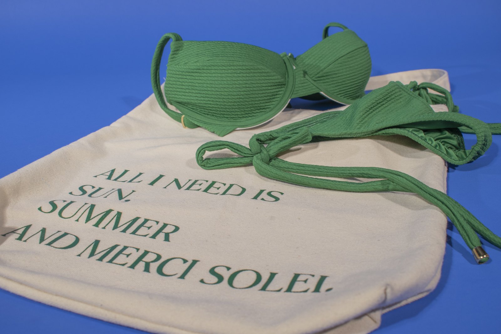 Green Merci Solei bikini and branded tote against blue